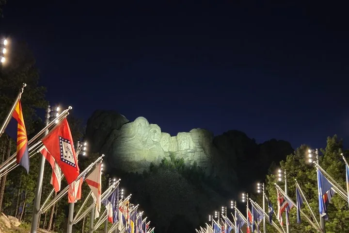 El Monte Rushmore iluminado de noche con una fila de banderas estatales al frente.
